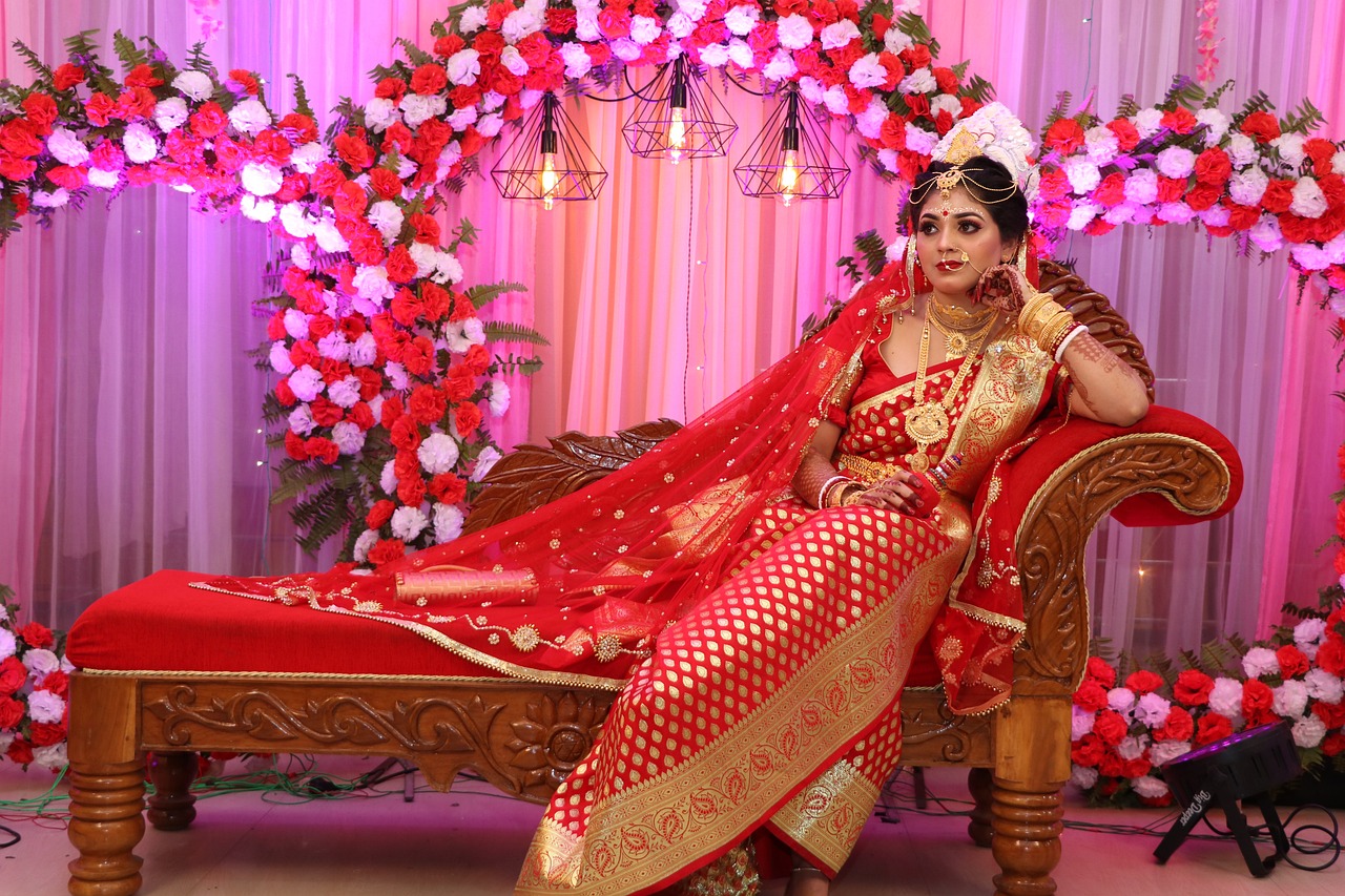 Traditional Indian bride in a red and gold saree seated on an ornate wooden couch, adorned with jewelry and vibrant floral decor at a wedding ceremony, captured by professional wedding photographer Suraj Verma.